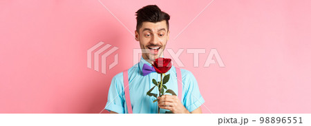 Valentines day and romance concept. Happy boyfriend looking at red rose while waiting for lover in bow-tie, standing on pink romantic background Valentines day and romance concept. Happy boyfriend looking at red rose while waiting for lover in bow-tie, standing on pink romantic background 98896551