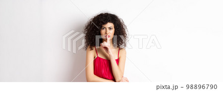 Angry elegant woman in red dress hushing and frowning, tell to be quiet, asking for silence, standing over white background 98896790