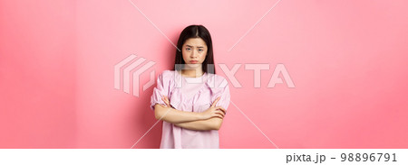 Silly asian girl feel unfair, cross arms on chest and sulking, frowning at camera, standing offended against pink background Silly asian girl feel unfair, cross arms on chest and sulking, frowning at camera, standing offended against pink background 98896791