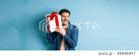 Valentines day. Smiling man guessing what inside gift box, got present from lover girlfriend, standing against blue background Valentines day. Smiling man guessing what inside gift box, got present from lover girlfriend, standing against blue background 98896847