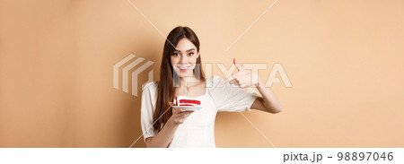 Cute girl pointing at birthday cake with candle and smiling excited, making bday wish, standing on beige background 98897046