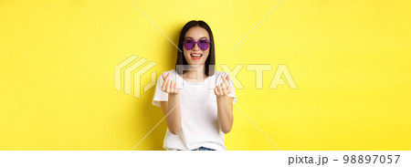Fashion and lifestyle concept. Attractive asian woman in heart-shape sunglasses, showing finger hearts and smiling happy at camera, standing over yellow background Fashion and lifestyle concept. Attractive asian woman in heart-shape sunglasses, showing finger hearts and smiling happy at camera, standing over yellow background 98897057