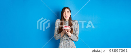 Dreamy birthday girl making wish with closed eyes and happy smile, blowing cake candle, standing on blue background Dreamy birthday girl making wish with closed eyes and happy smile, blowing cake candle, standing on blue background 98897061