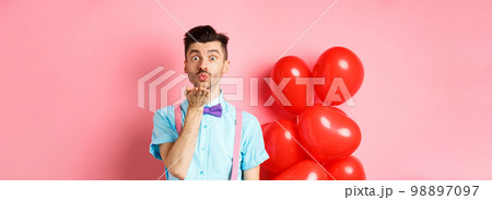 Valentines day concept. Attractive and funny man in bow-tie, blowing air kiss to lover, standing near pink background and red hearts balloons Valentines day concept. Attractive and funny man in bow-tie, blowing air kiss to lover, standing near pink background and red hearts balloons 98897097