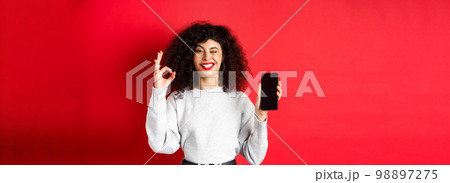 Attractive woman with smartphone, showing okay sign and empty phone sreen, recommending shopping app, standing against red background Attractive woman with smartphone, showing okay sign and empty phone sreen, recommending shopping app, standing against red background 98897275