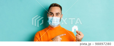 Real estate and coronavirus pandemic concept. Close-up of adult man in medical mask pointing at small paper house, standing over light blue background Real estate and coronavirus pandemic concept. Close-up of adult man in medical mask pointing at small paper house, standing over light blue background 98897280