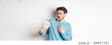 Surprised man holding money, looking amazed at camera, touching chest and gasping, standing in blue sweatshirt over white background Surprised man holding money, looking amazed at camera, touching chest and gasping, standing in blue sweatshirt over white background 98897281
