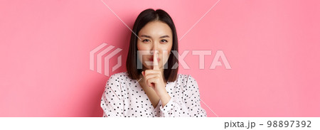 Close-up of mysterious asian woman hiding a secret, hushing and telling to keep quiet, standing over pink background Close-up of mysterious asian woman hiding a secret, hushing and telling to keep quiet, standing over pink background 98897392