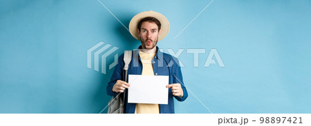 Excited tourist in straw hat, holding empty piece of paper and looking amused, going on summer travel, standing on blue background 98897421