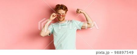 Confident young man with red hair, wearing summer sunglasses and t-shirt, showing strong and fit body muscles, flex biceps and staring cool at camera, pink background 98897549
