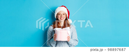 Winter holidays and Christmas Eve concept. Smiling redhead girl in sweater and Santa hat, holding New Year gift and looking at camera, standing against blue background 98897687
