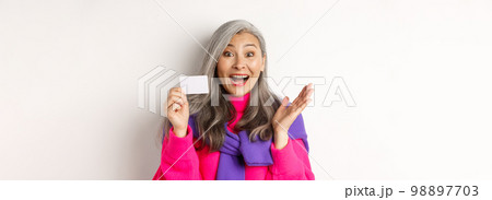 Shopping concept. Happy asian old aldy looking impressed and showing plastic credit card of her bank, standing over white background 98897703