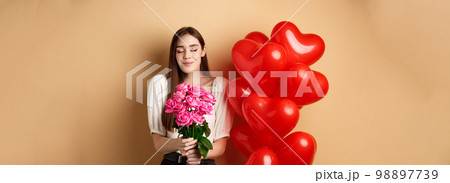 Romantic girlfriend smelling roses, receive flowers from lover on Valentines day, standing near red hearts balloons and smililng, beige background Romantic girlfriend smelling roses, receive flowers from lover on Valentines day, standing near red hearts balloons and smililng, beige background 98897739
