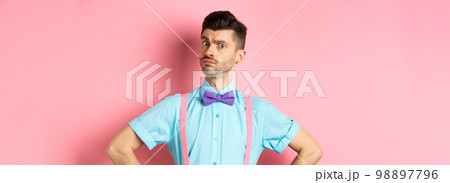 Image of young man with funny moustache and bow-tie standing in confident pose, staring with disbelief and doubts at camera, posing over pink background 98897796