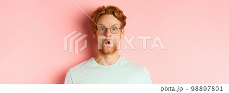 Close up of impressed redhead man in glasses saying wow, raising eyebrows surprised and staring at camera, standing over pink background 98897801