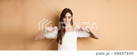 Young serious woman pointing at center, showing logo with confident face, standing in dress on beige background 98897840