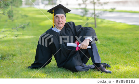 An elderly gray-haired man in a graduation gown sits on green grass. An elderly gray-haired man in a graduation gown sits on green grass. 98898182