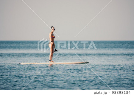 Sea woman sup. Silhouette of happy middle aged tanned woman in rainbow bikini, surfing on SUP board, confident paddling through water surface. Idyllic sunset. Active lifestyle at sea or river. Sea woman sup. Silhouette of happy middle aged tanned woman in rainbow bikini, surfing on SUP board, confident paddling through water surface. Idyllic sunset. Active lifestyle at sea or river. 98898184