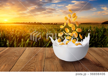 cornflakes falling in bowl with milk on table with green field on sunset as background 98898406