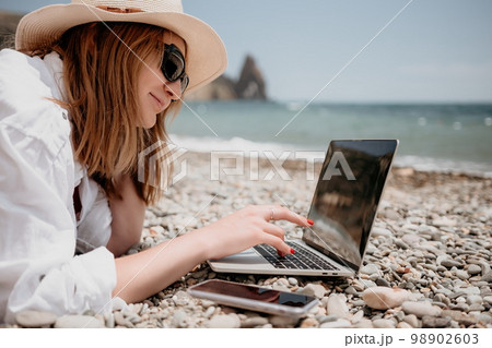Woman sea laptop. Business woman in yellow hat working on laptop by sea. Close up on hands of pretty lady typing on computer outdoors summer day. Freelance, digital nomad, travel and holidays concept. 98902603