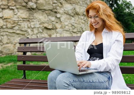 Young redhead smiling woman using her laptop sitting on the bench outdoors 98909812