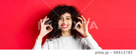 Close-up portrait of smiling woman with curly hair and red lips, showing okay gesture and looking satisfied, praise good product, standing on red background 98913785