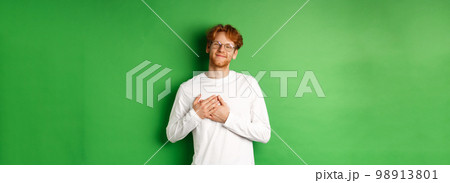 Handsome young man in glasses and white long-sleeve smiling, holding hands on heart with grateful face, thanking you, standing over green background 98913801