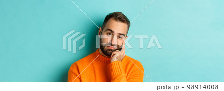 Close-up of unamused sad man looking at camera, pouting and leaning face on hand, standing over light blue background 98914008