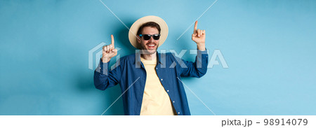 Cheerful caucasian guy in straw hat and sunglasses, dancing and having fun on vacation, standing over blue background. Concept of summer tourism 98914079