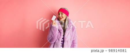 Shopping and fashion concept. Stylish asian senior woman in trendy clothes showing plastic credit card, smiling sassy, standing over pink background 98914081