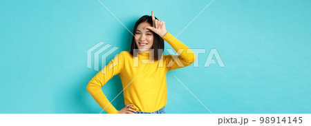 Sassy asian girl mocking lost team, showing loser sign on forehead and smiling pleased, being a winner, standing over blue background 98914145