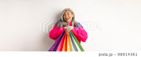 Fashionable senior asian woman shopaholic, hugging shopping bags and smiling joyful, buying with discounts, standing over white background 98914361