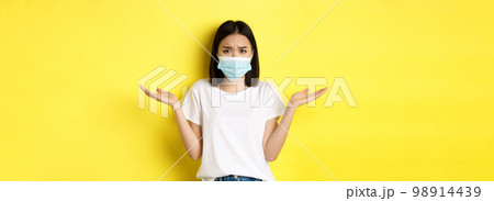 Covid-19, quarantine and social distancing concept. Confused asian woman in medical mask shrugging shoulders, spread hands sideways clueless, know nothing, yellow background Covid-19, quarantine and social distancing concept. Confused asian woman in medical mask shrugging shoulders, spread hands sideways clueless, know nothing, yellow background 98914439