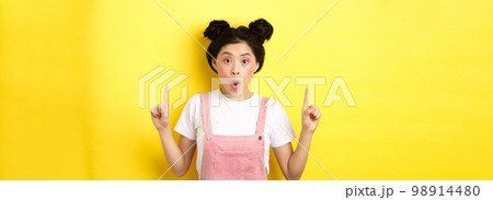 Excited asian female model with glamour makeup, pointing fingers up and say wow amazed, checking out promo deal, standing on yellow background Excited asian female model with glamour makeup, pointing fingers up and say wow amazed, checking out promo deal, standing on yellow background 98914480