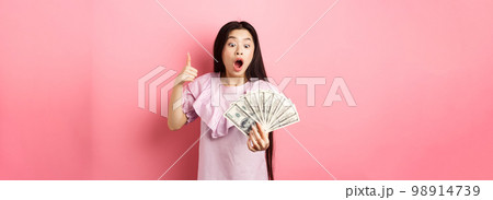 Excited teen girl holding big amount of money, showing dollar bills and thumbs up, standing amazed on pink background Excited teen girl holding big amount of money, showing dollar bills and thumbs up, standing amazed on pink background 98914739