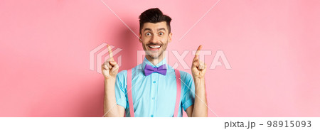 Portrait of cheerful young guy with moustache and bristle pointing fingers up, showing top logo, smiling happy at camera, standing on pink background 98915093