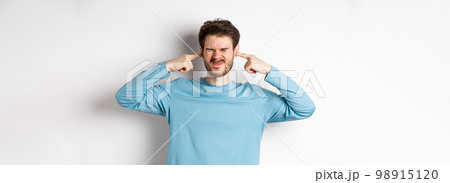 Annoyed young man plug ears from loud awful sound, being disturbed by bothering noise, grimacing displeased, standing on white background 98915120
