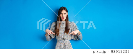Shocked and offended young woman in dress frowning, gasping and pointing fingers down at insulting promo, standing on blue background 98915235