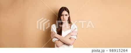 Offended woman hugging herself and frowning upset, feeling timid and defensive, standing on beige background 98915264