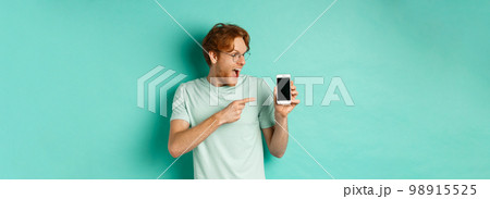 Check this out. Handsome redhead guy in glasses pointing finger at blank smartphone screen, showing online promotion, standing amazed over turquoise background Check this out. Handsome redhead guy in glasses pointing finger at blank smartphone screen, showing online promotion, standing amazed over turquoise background 98915525