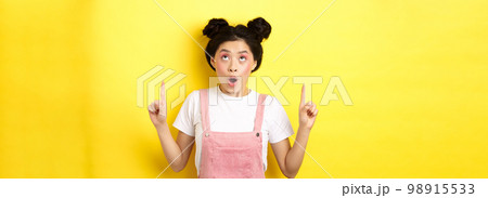 Excited asian teen student with glamour makeup, gasping amazed pointing fingers up, showing advertisement, yellow background 98915533