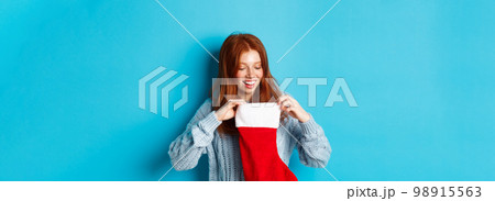 Winter holidays and gifts concept. Funny redhead girl looking inside Christmas stocking and smiling happy, receiving xmas present, standing against blue background Winter holidays and gifts concept. Funny redhead girl looking inside Christmas stocking and smiling happy, receiving xmas present, standing against blue background 98915563