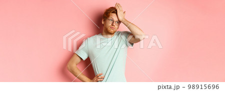 Tired young man with red hair, wearing glasses, looking annoyed and tensed, making facepalm gesture and exhale bothered, standing over pink background 98915696