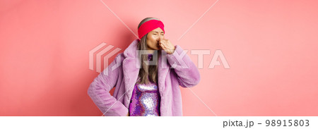 Portrait of stylish old asian woman in fashionable purple coat and dress, shut nose from bad smell, express disgust and aversion as something sink awfully, standing over pink background Portrait of stylish old asian woman in fashionable purple coat and dress, shut nose from bad smell, express disgust and aversion as something sink awfully, standing over pink background 98915803
