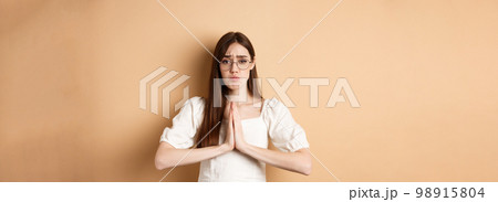 Sad girl in glasses begging for help, say please and looking cute, need favour, standing on beige background Sad girl in glasses begging for help, say please and looking cute, need favour, standing on beige background 98915804