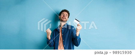 Happy and satisfied young man dancing with plastic credit card, fist pump and smile, standing on blue background Happy and satisfied young man dancing with plastic credit card, fist pump and smile, standing on blue background 98915930