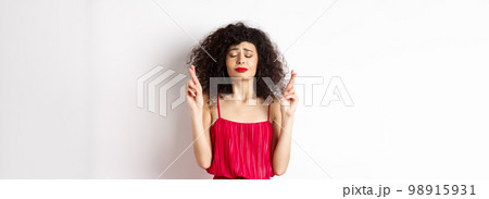 Hopeful woman in red dress making wish, praying with fingers crossed and closed eyes, pleading god, standing over white background Hopeful woman in red dress making wish, praying with fingers crossed and closed eyes, pleading god, standing over white background 98915931