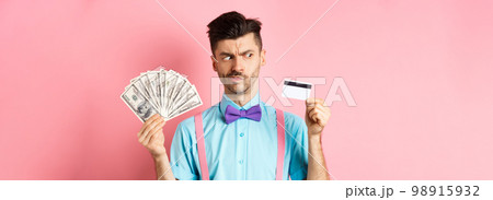 Doubtful and indecisive guy looking hesitant at plastic credit card, holding cash dollars, standing over pink background Doubtful and indecisive guy looking hesitant at plastic credit card, holding cash dollars, standing over pink background 98915932