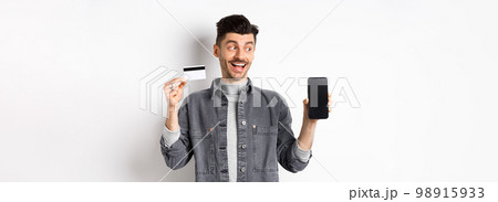 Handsome smiling man in denim jacket looking at empty smartphone screen and showing plastic credit card, good internet offer, standing against white background 98915933