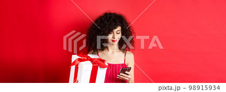 Valentines day and shopping. Beautiful caucasian woman in red dress, holding gift box from lover and using mobile phone, reading message on smartphone, studio background Valentines day and shopping. Beautiful caucasian woman in red dress, holding gift box from lover and using mobile phone, reading message on smartphone, studio background 98915934